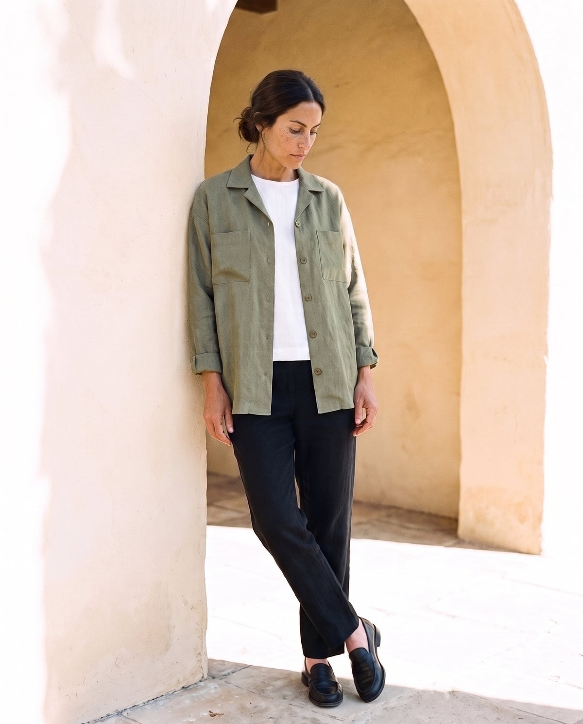 Rita Overshirt in moss, leaning against a sunlit wall