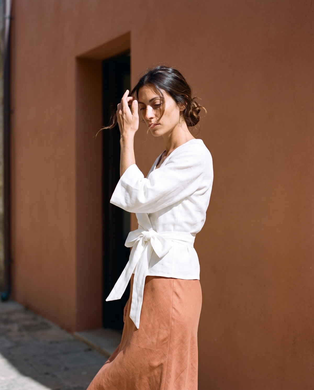 White tie front linen wrap top and clay bias cut slip skirt, pushing a strand of hair back in a shaded lane