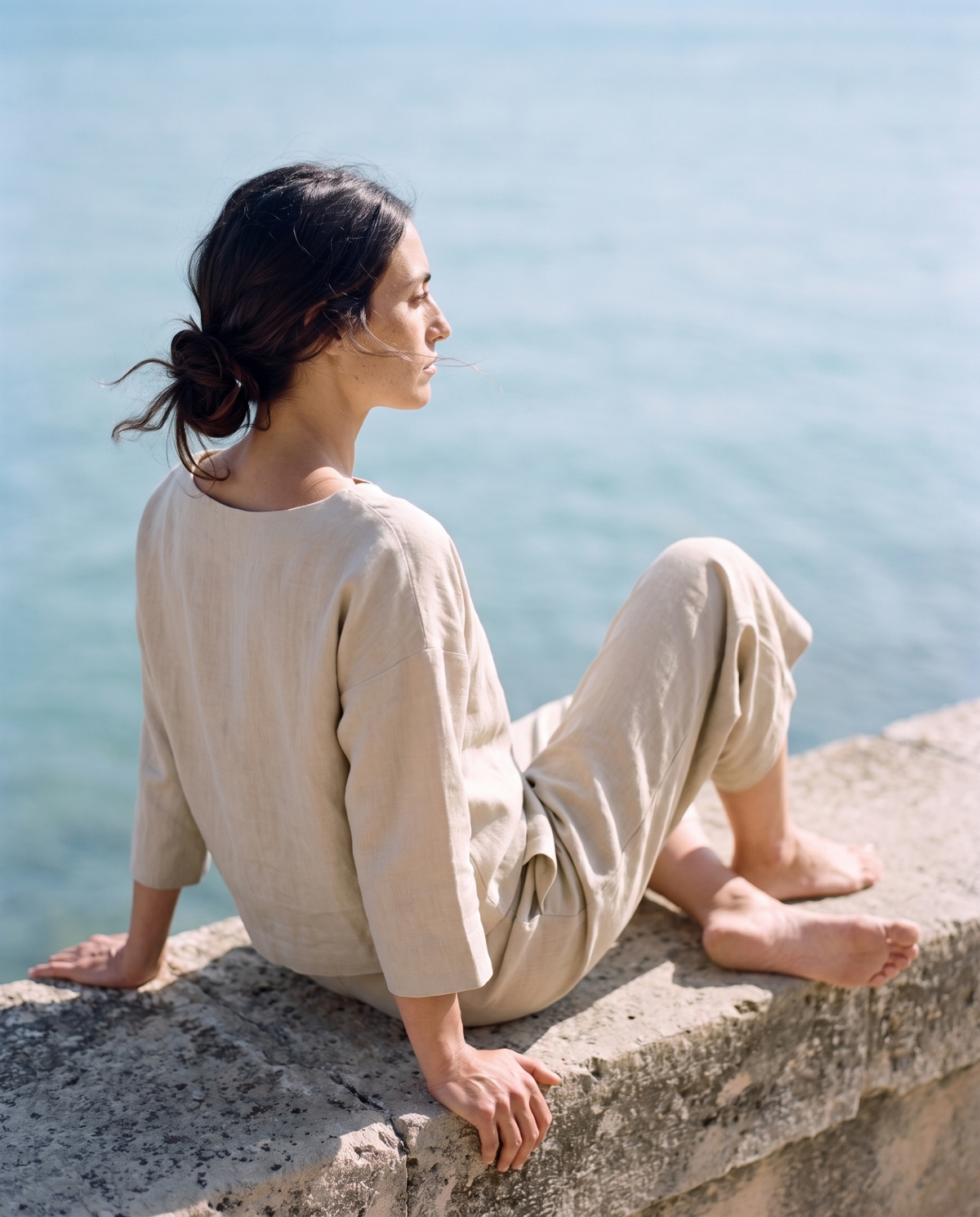 Natural boxy top and culottes set, sitting barefoot on a stone sea wall looking at the water