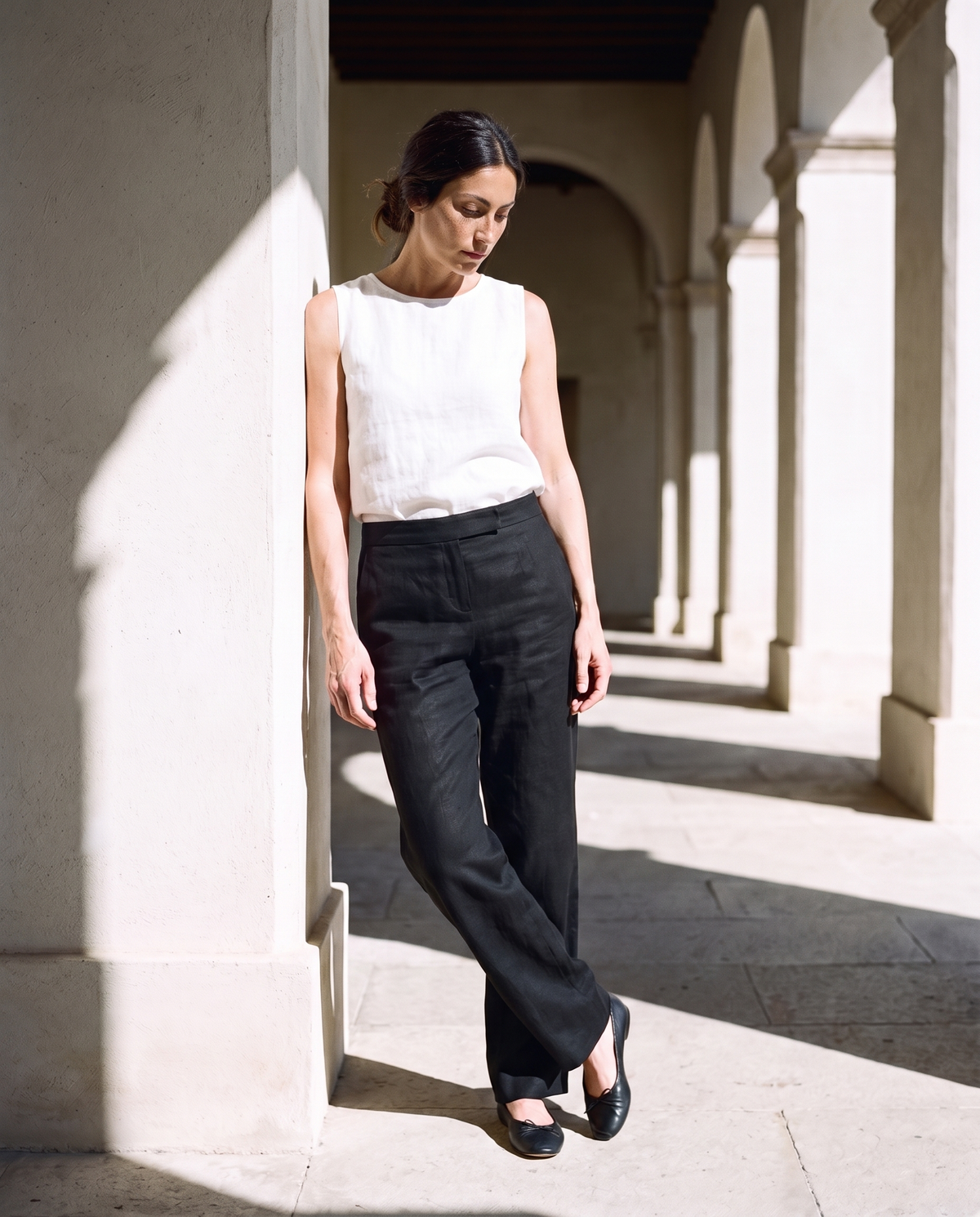 Nina Trousers in black, leaning against a sunlit wall
