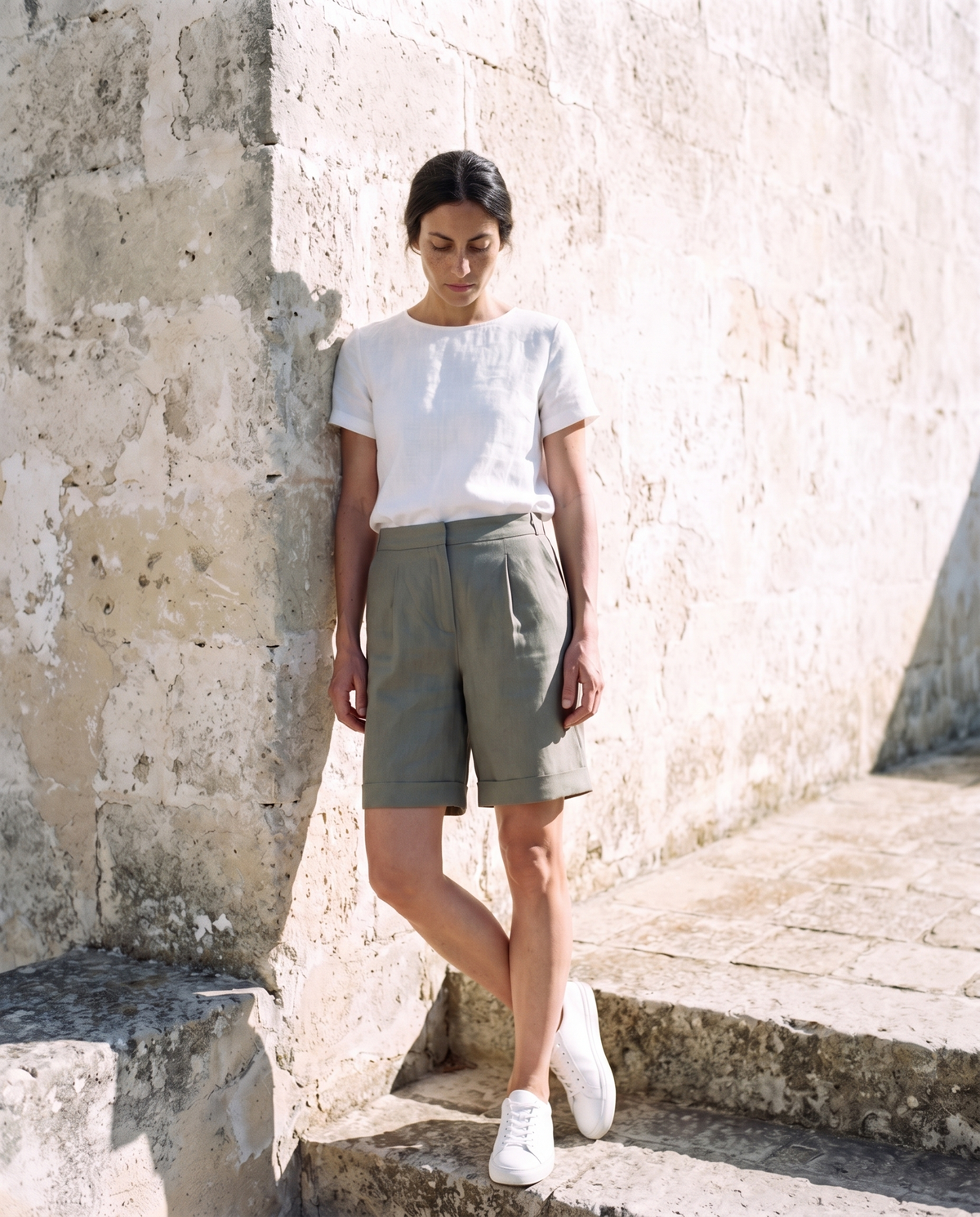 Rosa Shorts in moss, leaning against a sunlit wall