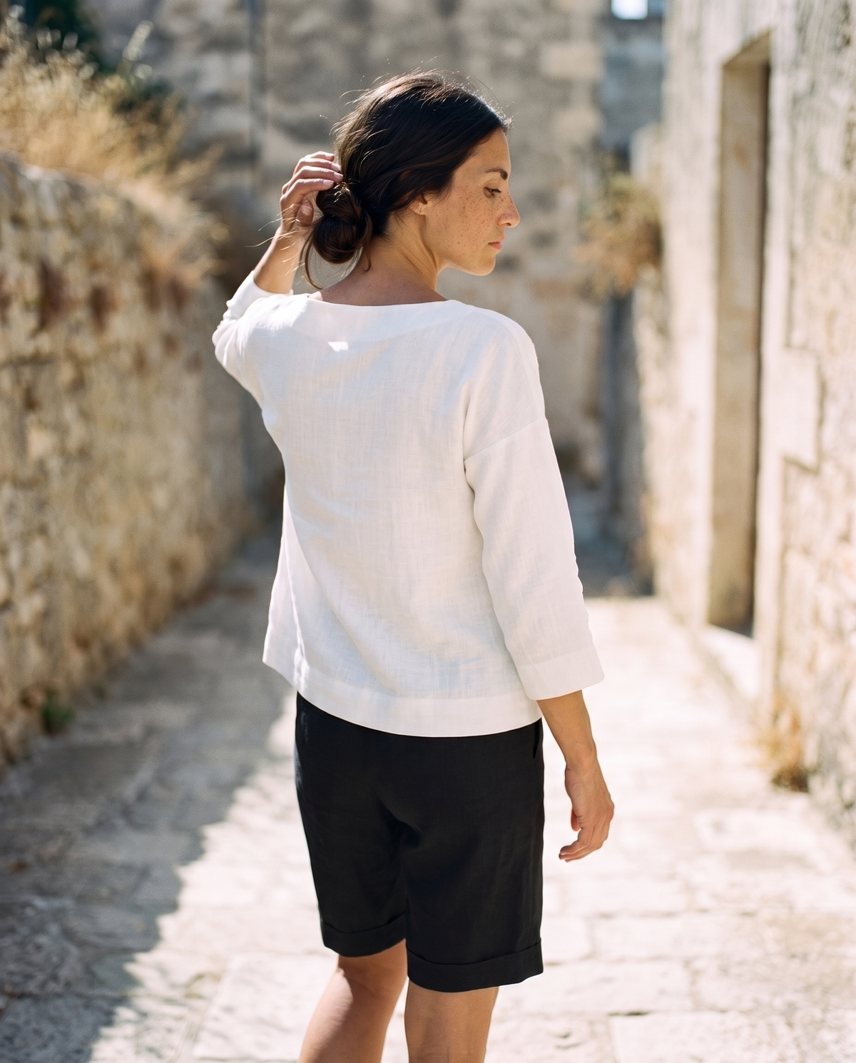 White boxy linen top and black Bermuda shorts, walking away down a lane lifting her hair off her neck