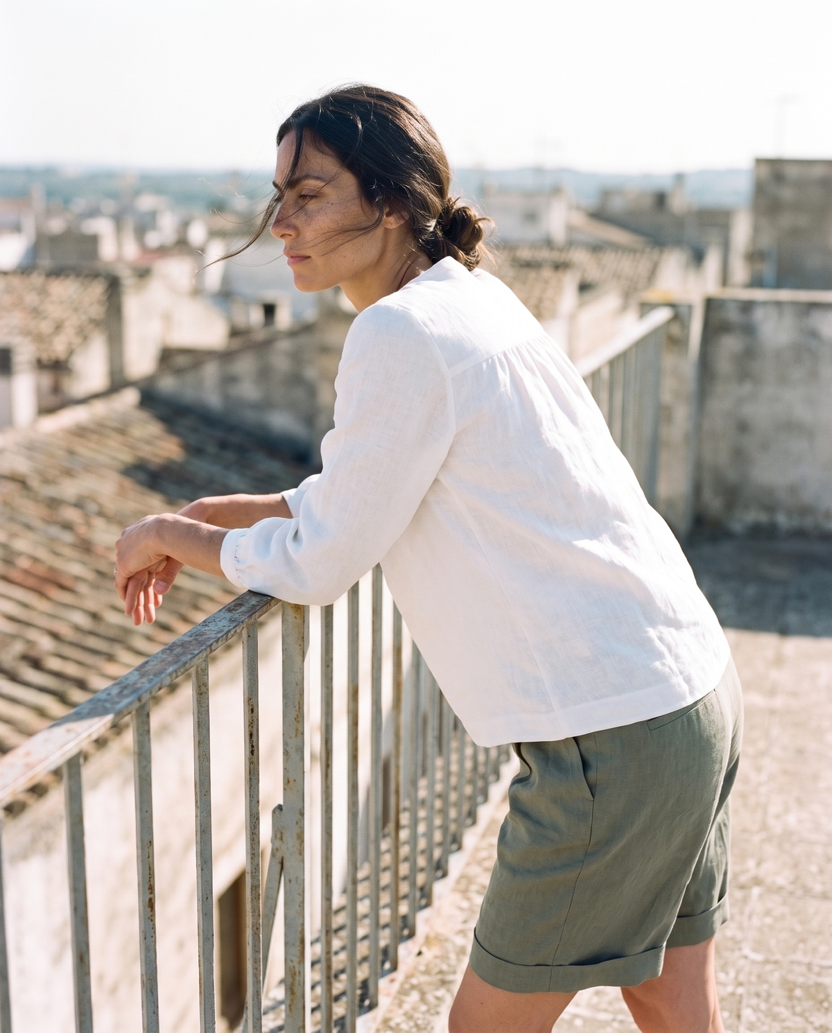 White gathered yoke linen blouse and moss Bermuda shorts, leaning on a balcony railing over the rooftops