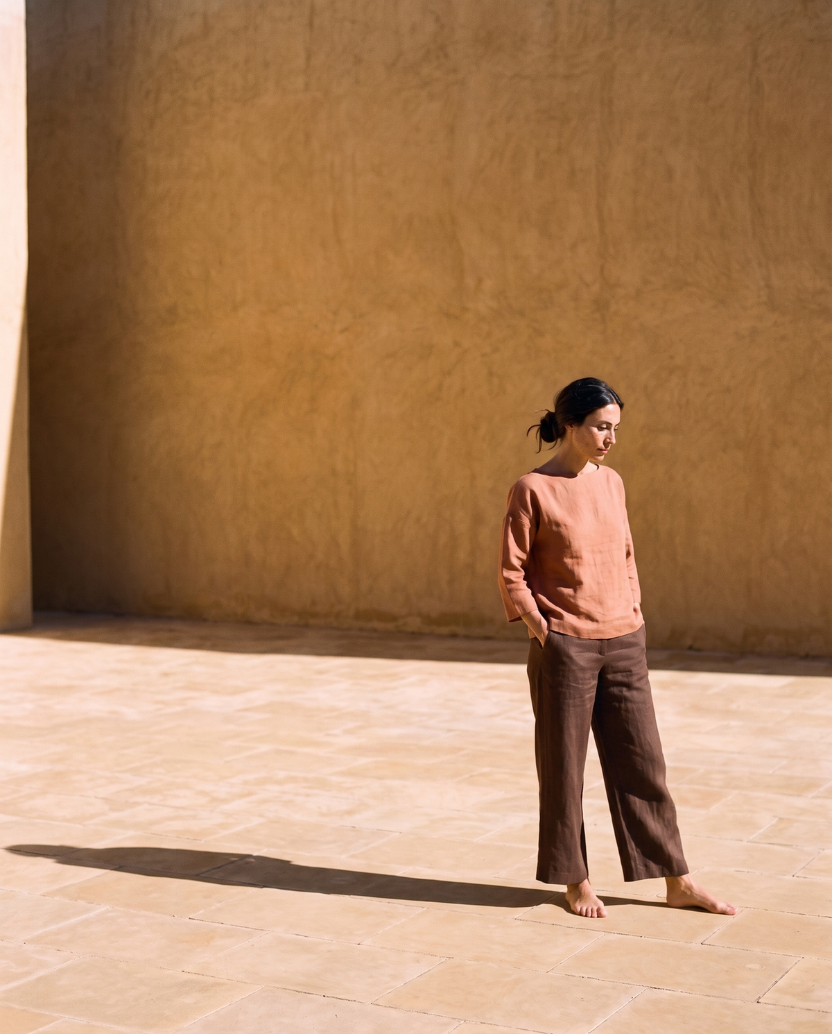 Clay boxy linen top and chocolate wide leg linen trousers, standing alone in a sunlit square with hands in pockets