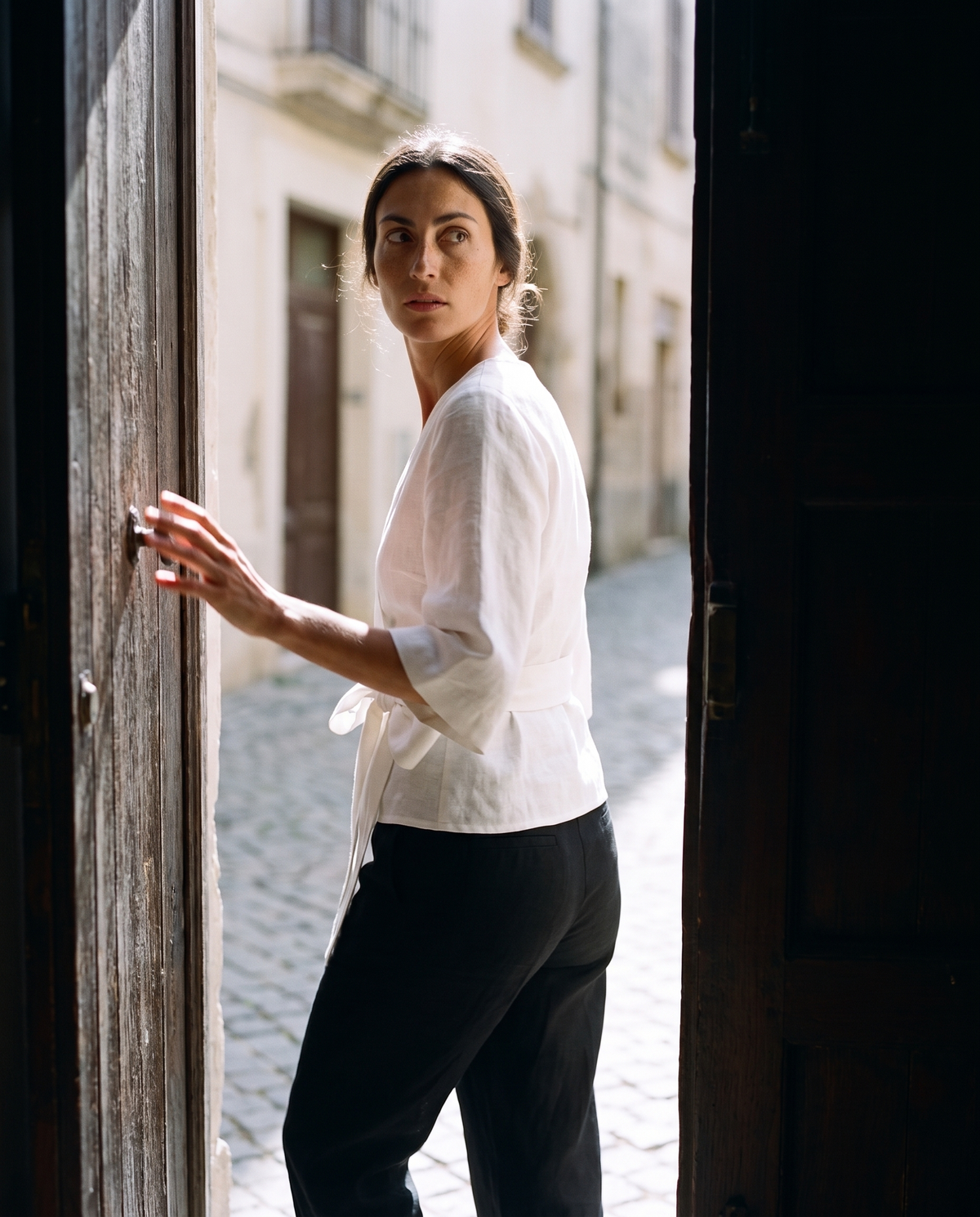 White tie front linen wrap top and black straight leg trousers, glancing back into the house from a doorway