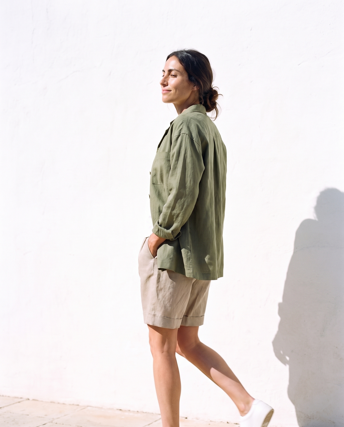 Moss linen overshirt over a white T-shirt with natural Bermuda shorts, walking past a whitewashed wall in hard sun