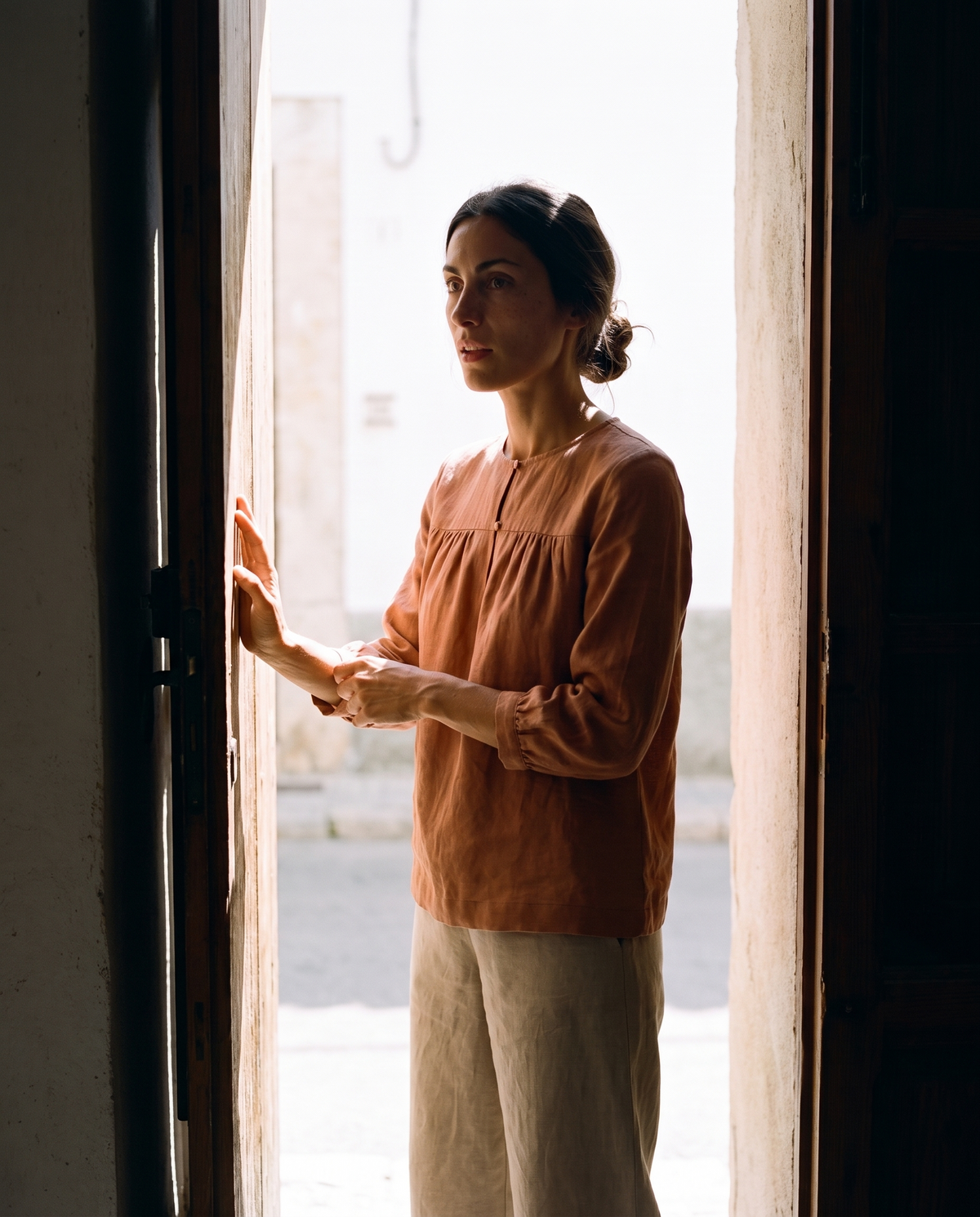 Clay gathered yoke linen blouse and natural culottes, standing in a doorway between a dark room and the bright street