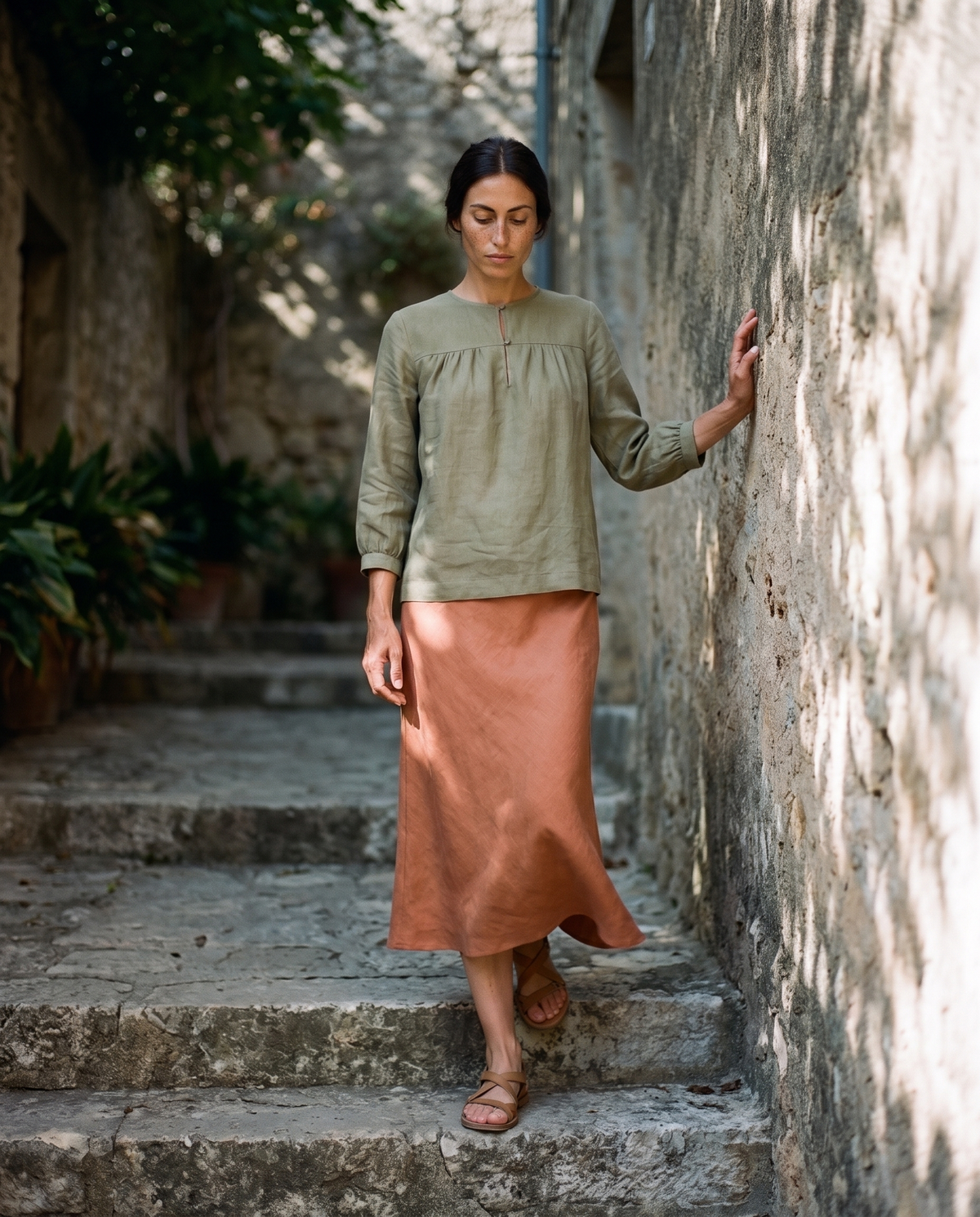 Moss gathered yoke linen blouse and clay slip skirt, taking the first step down a shaded stone stair