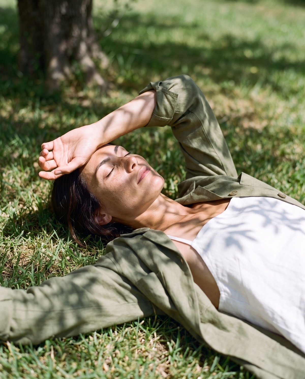 Moss linen overshirt over a white linen dress, lying on the grass in the shade with an arm over her face