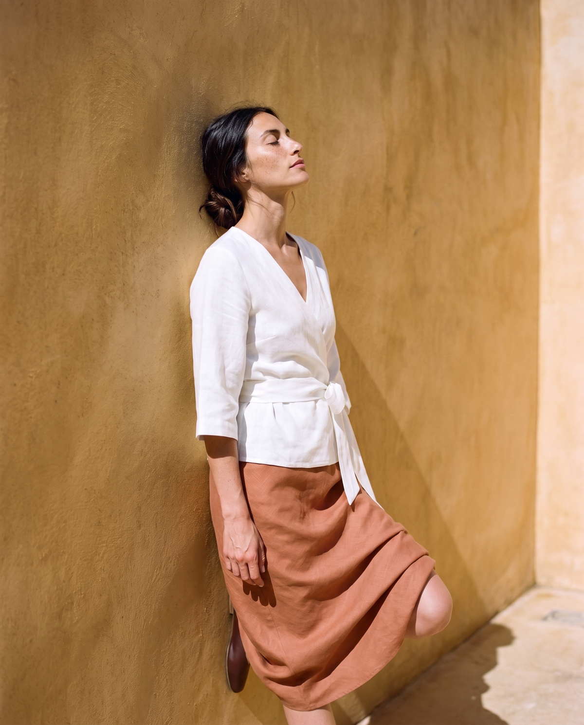 White tie front linen wrap top and clay slip skirt, leaning on a sunlit wall with eyes closed